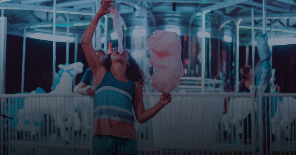 A girl joyfully holds a fluffy cotton candy in front of a colorful carousel, capturing a fun fair atmosphere.